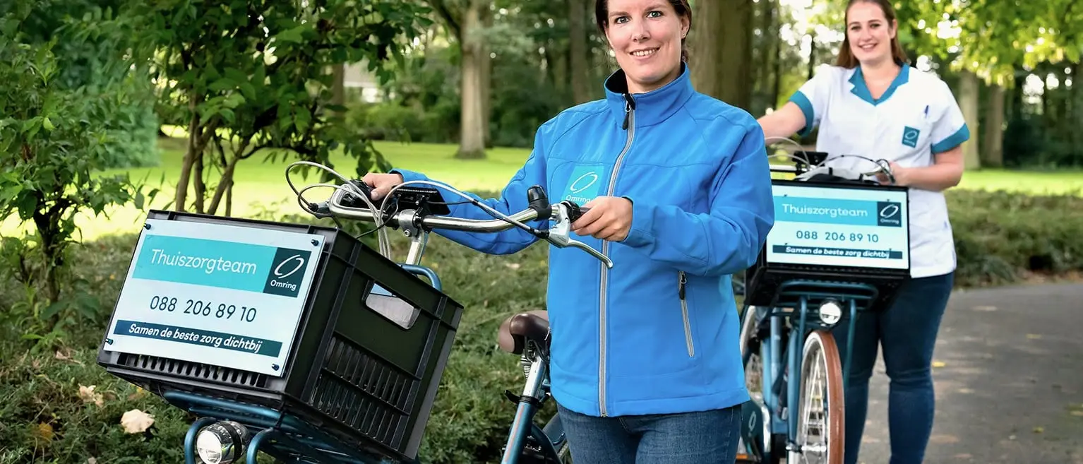 Twee Omring-thuiszorgcollega’s met hun fietsen, Thuiszorgteam-borden op de fietskratten